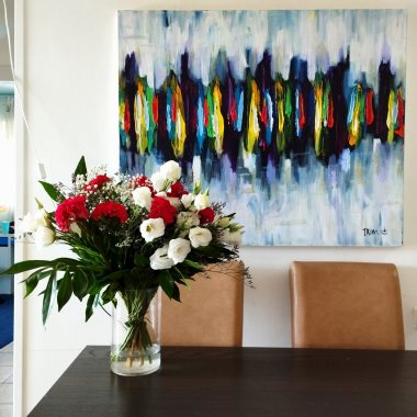 ציור מקורי: IMG_20220910_114028 מציג אגרטל עם פרחים אדומים ולבנים על שולחן כהה מול שני כיסאות חומים, עם ציור מופשט וצבעוני על קיר לבן בחדר אוכל מודרני. מוסיף צבע וייחודיות לכל חלל.