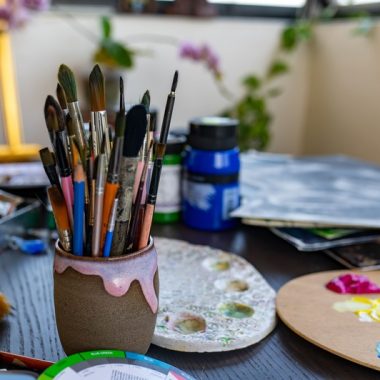 ציור מקורי: DSC_4333: כוס קרמיקה מלאה במכחולים מסוגים שונים מונחת על שולחן עמוס בצינורות צבע, פלטה, צנצנות וציוד אמנותי מסוגים שונים — מושלמת לשיעורי ציור או לשימוש בחבילת ציור בחדר מואר עם צמחים. מוסיף צבע וייחודיות לכל חלל.