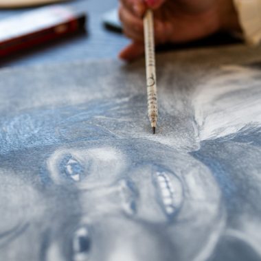 ציור מקורי: יד אוחזת ב-DSC_4342, ומציירת על נייר דיוקן מפורט של ילד מחייך עם עיניים ושיניים אקספרסיביות, בעוד שברקע נראים חומרי ציור מטושטשים מקורס רישום. מוסיף צבע וייחודיות לכל חלל.