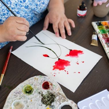 ציור מקורי: אדם בחולצה כחולה עם הדפס פרחוני משתמש ב-DSC_4622 כדי לצייר פרחים אדומים על נייר לבן בסדנת אמנות בוטנית. על השולחן הכהה מונחים חומרי אמנות: פלטת צבעים, סט צבעי מים, מכחולים וכד מים. מוסיף צבע וייחודיות לכל חלל.