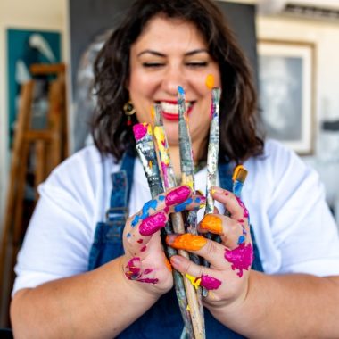 ציור מקורי: אישה מחייכת עם ידיים מלוכלכות בצבע מחזיקה מכחולים צבעוניים בסטודיו מואר, מוכנה לחוג ציור למבוגרים. היא לובשת חולצה לבנה וסינר כחול, ומזמינה אותך לנסות שיעור ניסיון של ציור ברמלה. | DSC_4819. מוסיף צבע וייחודיות לכל חלל.