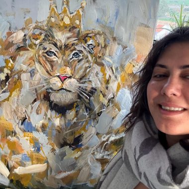 ציור מקורי: אישה עם שיער גלי וצעיף מחייכת ליד כן ציור שעליו מוצג 