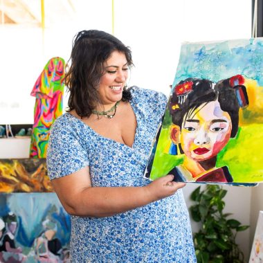 ציור מקורי: בסטודיו מואר, אישה בשמלה כחולה עם הדפס פרחוני מחייכת תוך שהיא מחזיקה את הציור המקורי 