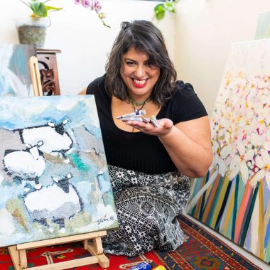 ציור מקורי: אישה מחייכת יושבת על שטיח בין ציורים צבעוניים, אוחזת בידה פסלון קטן של כבשה. לפניה נמצא il_fullxfull.7079202146_add9.jpg, ציור כבשה מקסים המוקף באמנות קיר פרחונית על בד קנבס לעיצוב בית חווה. מוסיף צבע וייחודיות לכל חלל.