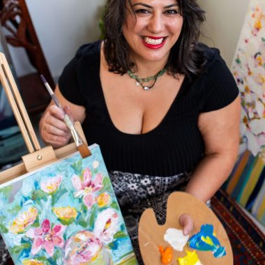 ציור מקורי: אישה מחייכת עם שיער כהה מחזיקה פלטה צבעים ומכחול ליד כן ציור שעליו מוצג ציור שמן פרחוני צבעוני בעבודת יד, מוקף בצמחים ויצירות אמנות, עם il_fullxfull.7079208522_p6io.jpg קנבס איטלקי. מוסיף צבע וייחודיות לכל חלל.