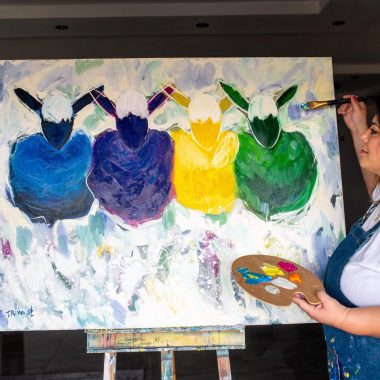 ציור מקורי: אישה בחלוק מציירת ציור פופ ארט מופשט ותוסס של ארבע דמויות חסרות פנים בצבעים כחול, סגול, צהוב וירוק על בד גדול שכותרתו il_fullxfull.7127078147_gsiz.jpg, כשהיא אוחזת בפלטה ומכחול בחלל פנימי מואר. מוסיף צבע וייחודיות לכל חלל.