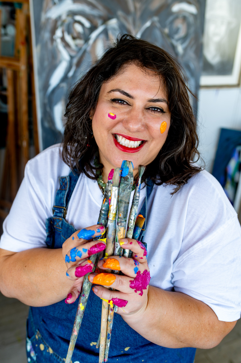 ציור מקורי: בתמונה DSC_4831, אישה מחייכת עם שיער כהה, לבושה בחולצה לבנה ובאוברול כחול, אוחזת בכמה מכחולים. ידיה ופניה מכוסות בכתמי צבע צבעוניים, וברקע ניתן לראות ציור ססגוני. מוסיף צבע וייחודיות לכל חלל.