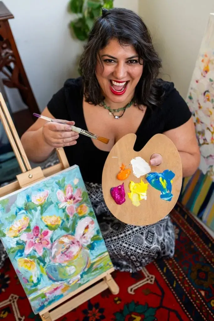 ציור מקורי: אישה מחייכת חיוך רחב כשהיא אוחזת במכחול ובפלטה ליד ציור פרחים על כן ציור. היא יושבת על שטיח מעוצב בחדר מלא חיים ואמנות. מוצר מוצג: DSC_4693. מוסיף צבע וייחודיות לכל חלל.