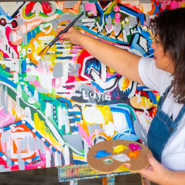 ציור מקורי: אישה בחולצה לבנה וסינר כחול מציירת במיומנות אמנות מופשטת על בד עם פלטת צבעים ססגונית. המילה LOVE מודגשת ביצירת האמנות המודרנית שלה, המוצגת במוצר 