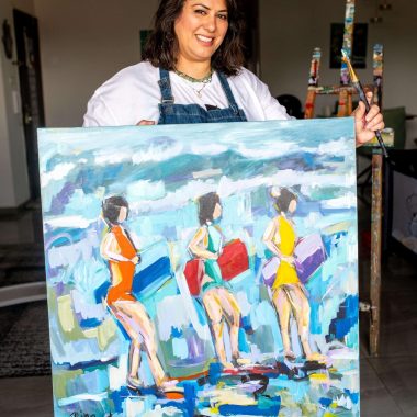 ציור מקורי: אישה מחייכת בתוך הבית, לבושה בסינר ומחזיקה מכחול, תוך שהיא מציגה את il_fullxfull.7079129548_157q.jpg — בד צבעוני המציג שלוש נשים בבגדי ים על החוף עם ציוד אמנותי ברקע. מוסיף צבע וייחודיות לכל חלל.