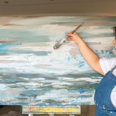 ציור מקורי: אישה בחלוק כחול וחולצה לבנה מחייכת בזמן שהיא מציירת על בד גדול ומופשט בצבעים כחול, לבן ואפור בחדר מודרני ומואר, עם יצירת אמנות על הקיר בשם 