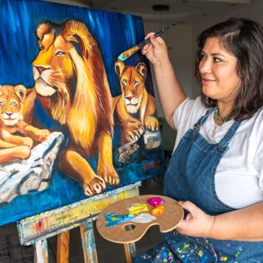 ציור מקורי: אישה בחלוק ג'ינס מציירת את il_fullxfull.7127031737_cpse.jpg, ציור שמן מלא חיים של משפחת אריות הכוללת אריה ושלושה גורים. מחייכת עם לוח הצבעים שלה, היא יוצרת את המתנה המושלמת לאוהבי בעלי חיים. מוסיף צבע וייחודיות לכל חלל.