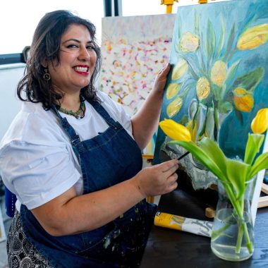 ציור מקורי: אישה מחייכת בזמן שהיא מציירת צבעונים על בד איטלקי בסטודיו לאמנות מואר, בהשראת אגרטל פרחים וחומרי ציור, בזמן שהיא יוצרת את הציור בשמן 